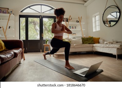 Healthy black African American woman with afro watching video tutorials on laptop while exercising with hands joined on yoga mat at home - woman doing warrior pose
