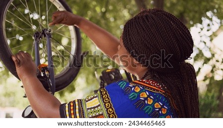 Healthy black woman mending her own bicycle using specialized equipment from toolkit in yard. Dolly zoom-in shot. Female african american cyclist repairing front bike wheel outdoors.