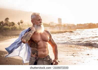 In a healthy body, healthy mind. Senior man with white stylish beard and tattoos walking on the beach at sunset. Summer vacation . Elderly people healthy lifestyle on a retirement concept.