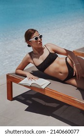 Healthy Body Young Beautiful Asian Multiracial Woman Wearing Sunglasses Lying Down On Lounge Chair Enjoying Sun Tanning Sunbathing In Swimwear Reading Book By Pool Side On Summer Vacation.