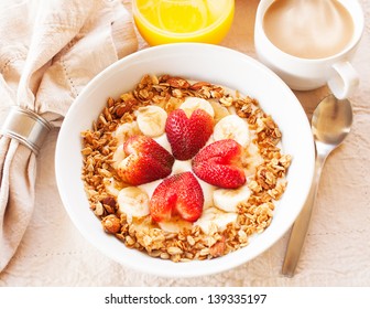 A healthy breakfast consisting of vanilla greek yogurt topped with granola, sliced bananas and strawberries shaped like hearts. Orange juice and coffee with steam are also present.