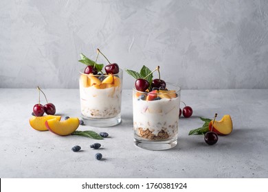 Healthy breakfast in a glass with fresh fruits: pomegranate, cherry, nectarines, honeysuckle, yogurt and granola on a grey background. Shallow depth of field with selective focus