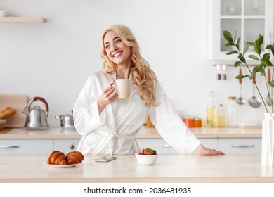 Healthy breakfast at home, good morning and delicious coffee, rest and enjoy weekend and vacation. Cheerful beautiful young caucasian female in bathrobe drinking coffee in minimalist kitchen interior - Powered by Shutterstock