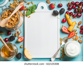 Healthy Breakfast Ingredients. Oat Granola In Open Glass Jar, Yogurt, Fruit, Berries, Honey And Mint On Blue Background With White Ceramic Board In Center, Top View, Copy Space