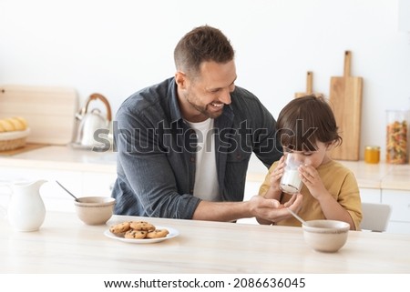 Healthy breakfast for kids. Positive loving father giving glass of fresh milk to his little son, asirabble boy enjoying tasty drink, sitting together at home kitchen