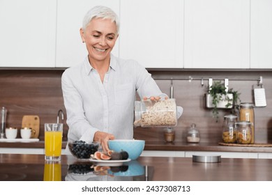 Healthy breakfast in the morning concept. Caucasian middle-aged mature woman preparing oat flakes porridge with fruits at home kitchen. Dieting and slimming - Powered by Shutterstock