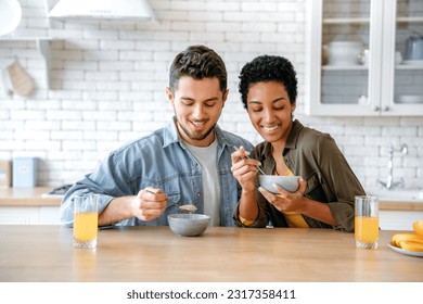 Healthy breakfast. Weekend morning. Happy young family couple, caucasian man and african american woman, having breakfast at cozy home kitchen with healthy porridge, juice and fruits, smiling - Powered by Shutterstock