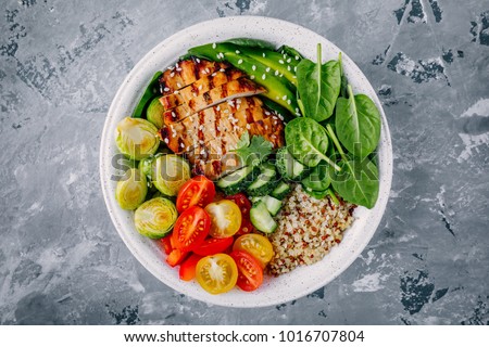 Healthy buddha bowl lunch with grilled chicken, quinoa, spinach, avocado, brussels sprouts, tomatoes, cucumbers  on dark gray background. Top view.
