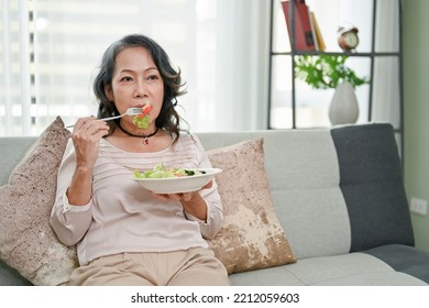 Healthy And Calm 60s Retired Asian Woman In Casual Clothes Spending Her Weekend At Home, Eating Healthy Salad While Watching TV In Her Living Room.