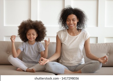 Healthy calm african american mother teaching meditation sit with cute small child daughter on sofa, mindful happy black mom and kid girl doing yoga exercises relaxing together in lotus pose at home - Powered by Shutterstock