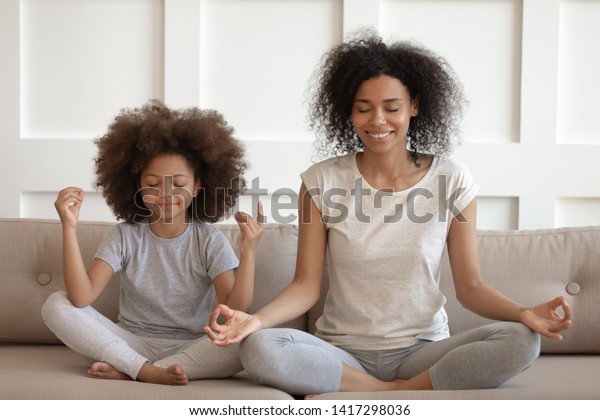 Healthy calm african american mother teaching\
meditation sit with cute small child daughter on sofa, mindful\
happy black mom and kid girl doing yoga exercises relaxing together\
in lotus pose at home