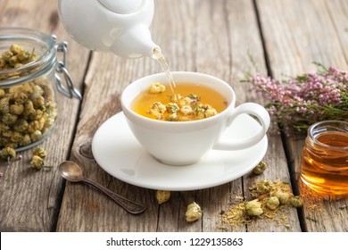 Healthy chamomile tea poured into white cup. Teapot, small honey jar,  heather bunch and glass jar of daisy medicinal herbs.
