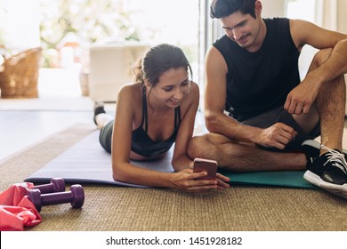 Healthy couple doing workout at home. Woman lying on floor showing her mobile phone to her boyfriend sitting by and having conversation after workout.