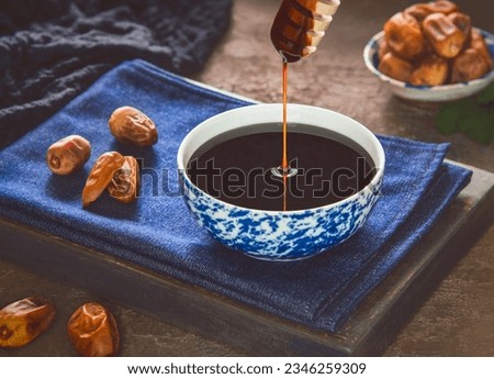 Healthy date syrup with whole and dry pitted dates. Delicious date syrup flows from a wooden honey dipper into a small bowl. It is natural sweetener made with dates.