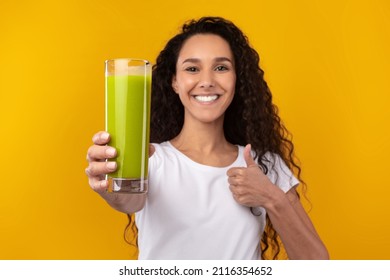 Healthy Detox Concept. Smiling young woman holding green smothie drink, giving offering glass close to camera, showing thumbs up gesture isolated on yellow orange studio wall, selective focus on hand - Powered by Shutterstock