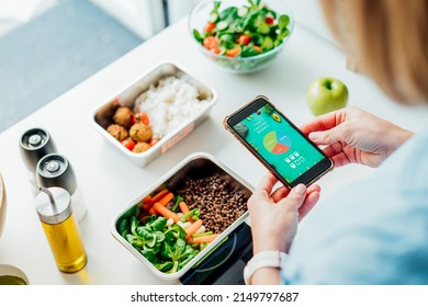 Healthy diet plan for weight loss, daily ready meal menu. Woman using meal tracker app on phone while weighing lunch box cooked in advance on kitchen scale. Balanced portion with dish. Pre-cooking - Shutterstock ID 2149797687