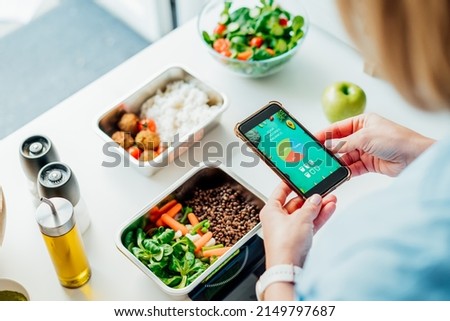Healthy diet plan for weight loss, daily ready meal menu. Woman using meal tracker app on phone while weighing lunch box cooked in advance on kitchen scale. Balanced portion with dish. Pre-cooking