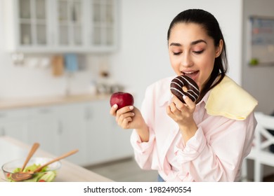 Healthy Diet vs Junk Food Concept. Portrait of young woman holding red apple fruit and doughnut in her hands, biting and eating sweet dessert. Smiling female choosing unhealthy meal, addicted to sugar - Powered by Shutterstock