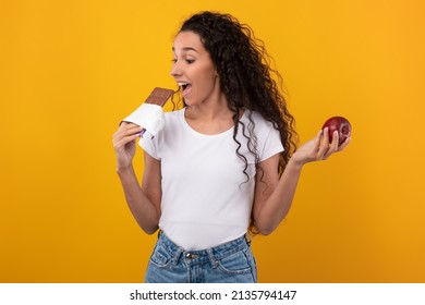 Healthy Diet vs Junk Food Concept. Portrait of young woman holding red apple fruit and chocolate in hands, biting and eating sweet dessert. Smiling female choosing unhealthy meal, addicted to sugar