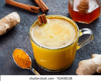 Healthy drink golden turmeric latte in glass cup.Gold milk with turmeric,ginger root,cinnamon sticks,dried turmeric powder and honey over black cement background.Detox turmeric tea and ingredients.