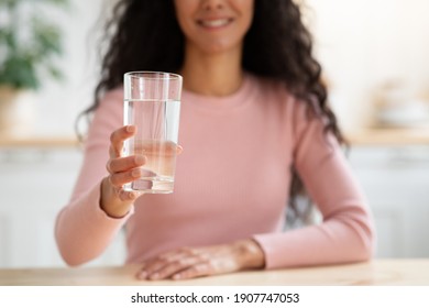 Healthy Drink. Unrecognizable Smiling Lady Offering Glass With Mineral Water At Camera While Sitting At Table In Kitchen, Brunette Woman Drinking Aqua At Home, Closeup Shot With Selective Focus