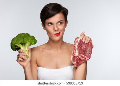 Healthy Eating. Beautiful Woman With Red Lips Choosing Between Raw Meat And Broccoli, Thinking Whether To Eat Vegetable Or Meat. Girl Holding Food Products In Hands. Diet Concept. High Resolution