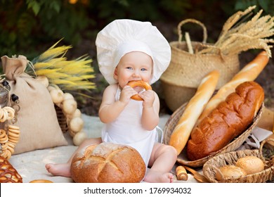 Healthy Eating Concept. Happy Boy Is Cooking On Nature On A Sunny Summer Day. Toddler Baker On A Picnic Eats Bread And Bagels In A White Apron And Hat