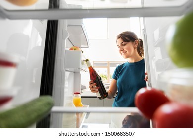 Healthy Eating, Food And Diet Concept - Happy Woman Taking Orange Wine Bottle From Fridge At Home Kitchen