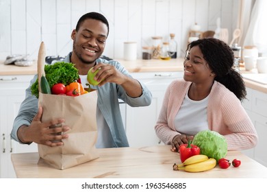 Healthy Eating. Happy young african american spouses unpacking paper bags with groceries at home, black couple sitting at table in kitchen and taking out vegetables and fruits after food shopping - Powered by Shutterstock