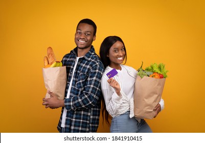 Healthy Eating, Modern Lifestyle And Grocery Shopping. Cheerful Millennial African American Couple In Casual Hold Paper Bags With Food And Show Credit Card Isolated On Yellow Background, Studio Shot