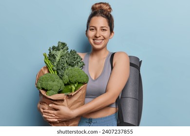 Healthy eating and sporty lifestyle concept. Cheerful active European woman embraces paper bag full of fresh green vegetables carries rubber karemat on shoulder isolated over blue background.