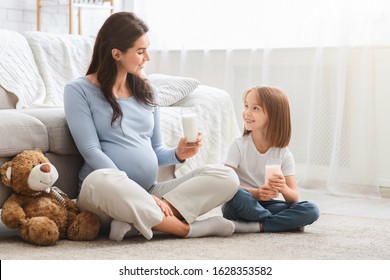 Healthy Expecting Young Mom And Little Cute Daughter Having Glass Of Milk, Sitting On Floor At Living Room