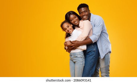 Healthy Family Relationship. Portrait of African American dad and mommy embracing smiling teen daughter standing isolated on yellow orange studio background, banner, panorama, free copy space
