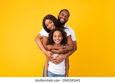 Healthy Family Relationship. Portrait Of Cheerful African American Man Hugging His Wife And Daughter From Behind Back Standing Isolated On Yellow Orange Studio Wall, Posing Looking At Camera