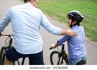 Healthy Family Relationship. Rear back view portrait of smiling father and son riding bicycles down the street in public park, man tapping and patting boy on the shoulder, teaching and showing support