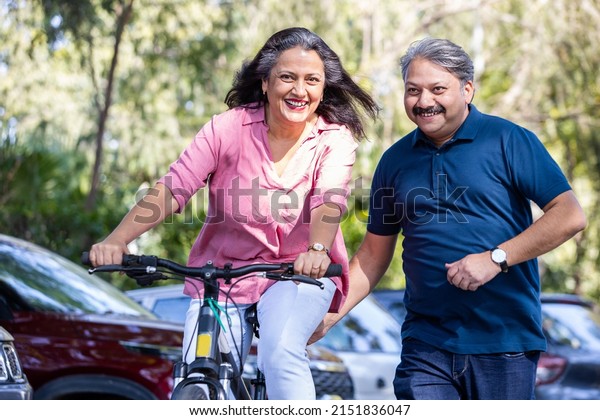 Healthy and fit old  Indian couple riding bicycle in the\
park summer, active old age people and lifestyle. Elderly woman\
learn to ride cycle with man. retired people having enjoy life.\
