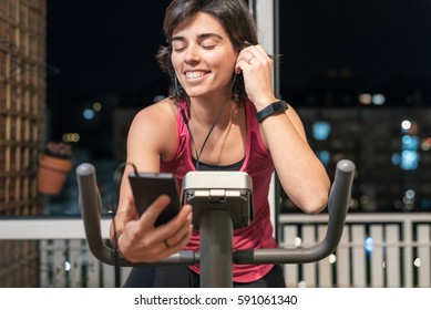 Healthy Fit Smiling Woman Training At Home On Exercise Static Bike During Workout Listening Playlist Music With Earphones Connected To Smart Phone For Motivation. Female Health Weekly Habits App