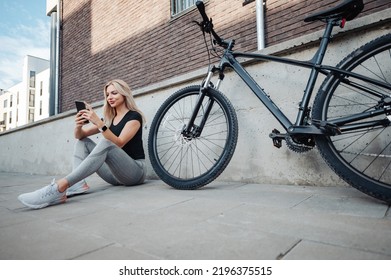 Healthy And Fit Woman In Sportswear Using Modern Smartphone While Sitting On Street Near Black Bike. Female Cyclist Taking Break During Outdoors Training With Modern Gadget In Hands.