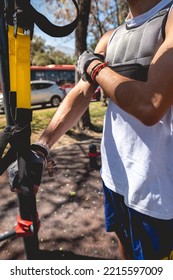 Healthy And Fit Young Latin Man With Weight Vest, Workout Gloves And Blue Shorts Performing Exercises With Elastic Bands On Bars In A Workout Park In Sunny Day