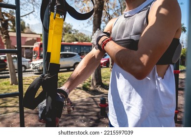 Healthy And Fit Young Latin Man With Weight Vest, Workout Gloves And Blue Shorts Performing Exercises With Elastic Bands On Bars In A Workout Park In Sunny Day