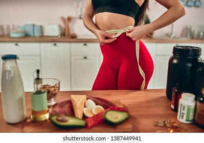 Healthy Food Concept. Cropped Image Of Attractive Sporty Woman Standing On Kitchen With Centimeter While Measuring Her Waist. Choice Between Healthy Food Full Of Protein And Sport Supplements. Sport