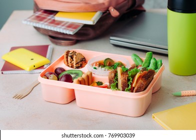 Healthy Food Concept: Open Lunch Box With Sandwiches, Vegetable Salad, Fresh Grape And Kiwi, Rice And Nuts On The Table Near Backpack, Laptop And Thermo Mug; Selective Focus