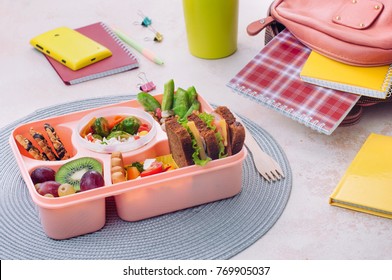 Healthy Food Concept: Open Lunch Box With Sandwiches, Vegetable Salad, Fresh Grape And Kiwi, Rice With Grilled Brussels Sprouts And Nuts On The Table Near School Backpack; Selective Focus