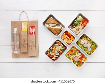 Healthy food delivery. Take away for diet. Fitness nutrition, vegetables, meat and fruits in foil boxes, cutlery, water and brown paper package. Top view, flat lay at white wood with copy space