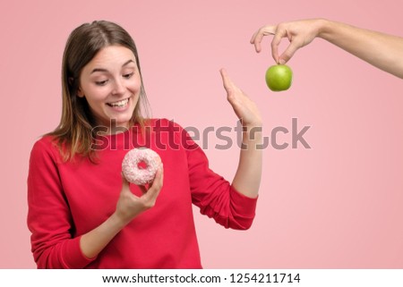 Healthy food and diet concept. Beautiful young woman in red clothes choosing between fruits and sweets standing on pink background. She prefer a delicious donut instead of green apple