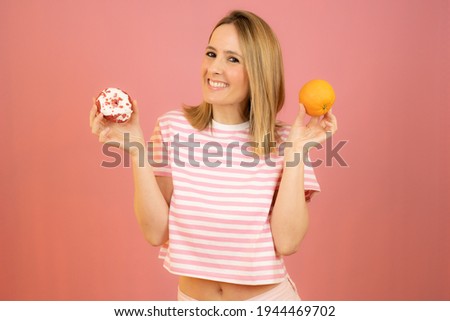 Healthy food and diet concept. Beautiful young woman in striped t-shirt choosing between fruits and sweets standing over pink background.