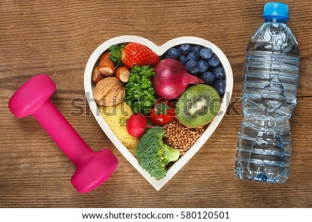 Healthy food in heart shaped bowl, bottle of water and dumbbell. Food such as blueberries, red onion, strawberry, parsley leaves, hazelnuts, walnut, tomato. kiwi, millet, buckwheat, radish, broccoli.
