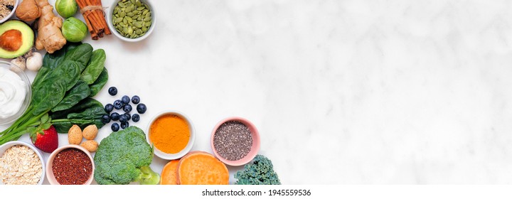 Healthy food ingredients. Top view corner border on a white marble banner background. Copy space. Super food concept with green vegetables, berries, whole grains, seeds, spices and nutritious items.