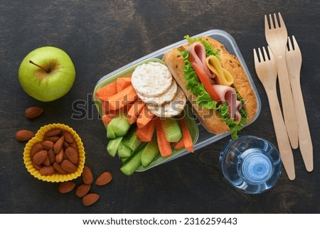 Healthy food for school lunch concept. Healthy bread, meat sandwich with cheese, apple, fresh cucumber, carrots, nuts in container, water and yogurt on dark background. Back to school concept.