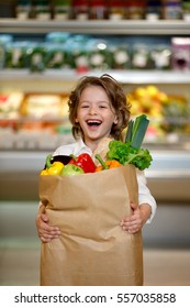 Healthy food for young family with kids. Portrait of smiling little child holding paper shopping bag full of fresh vegetables. Boy standing at grocery store or supermarket